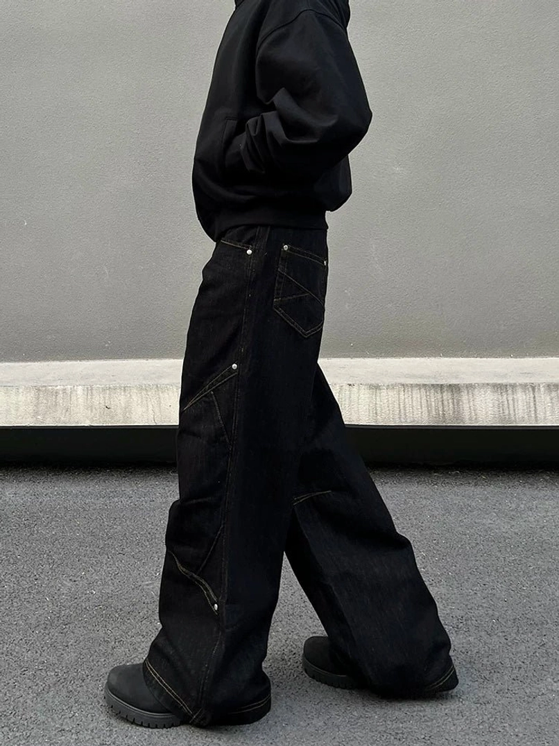 Side profile of black orbit panel streetwear jeans showing geometric thigh stitching, loose baggy silhouette, and long raw-style hem