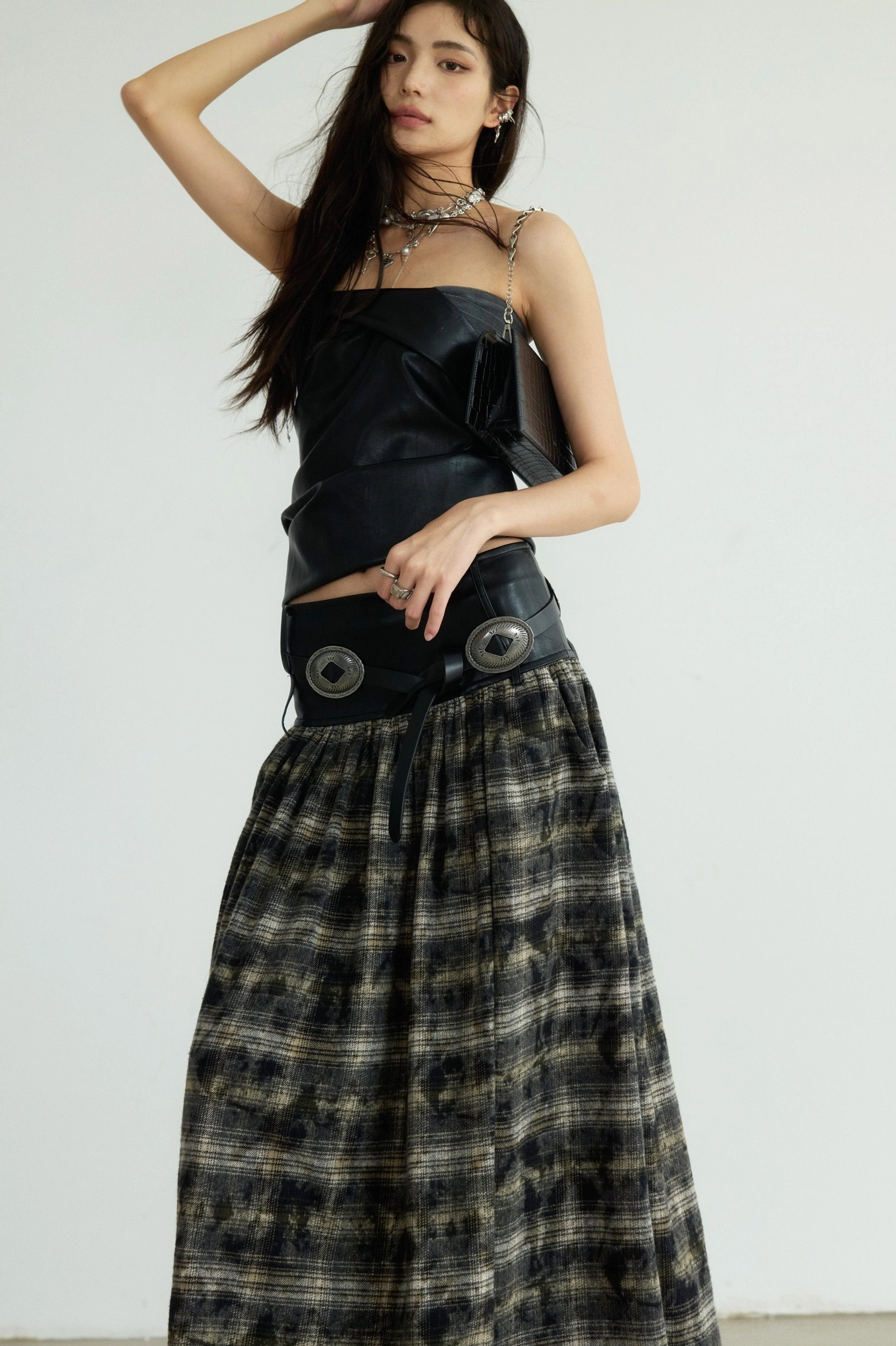 Side three-quarter view of a sculpted black origami bustier top with strapless neckline, fitted waist, faux leather finish, plaid maxi skirt, and chain bag