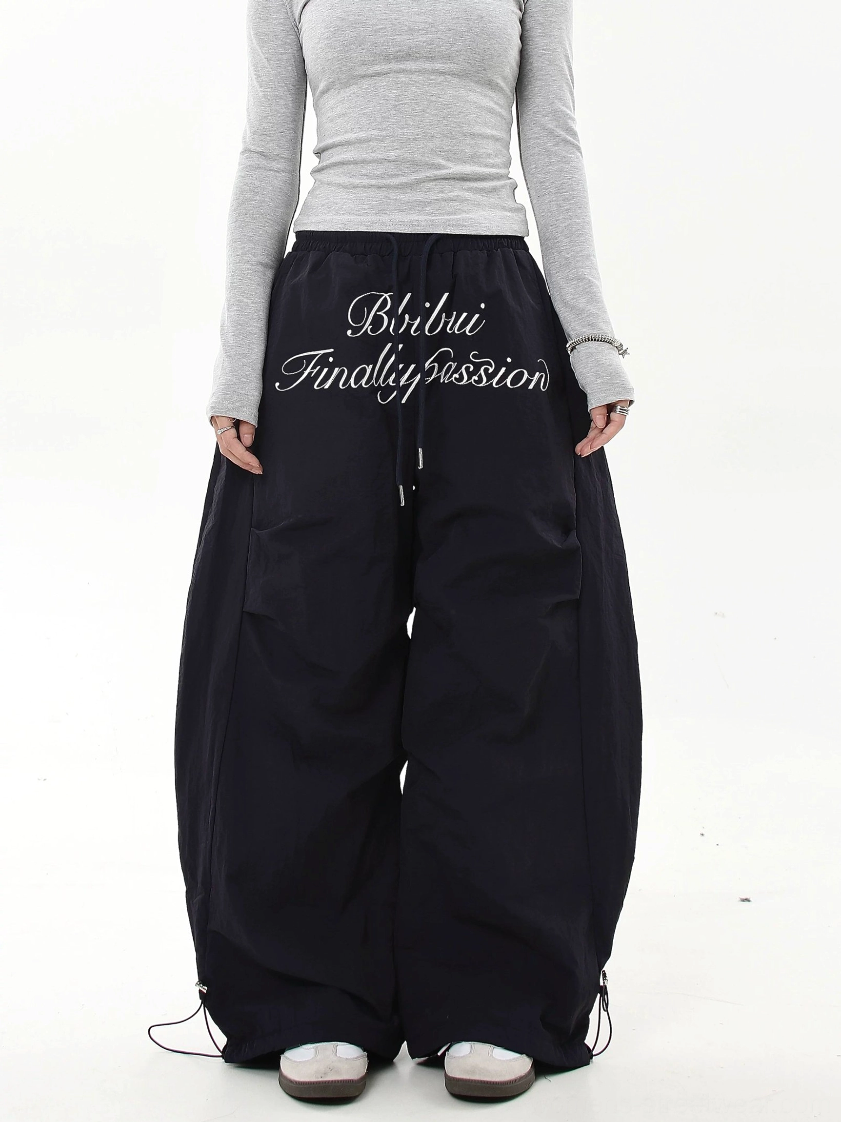 Black oversized baggy parachute cargo pants with white cursive embroidery, adjustable waist and ultra-wide legs styled for Korean streetwear