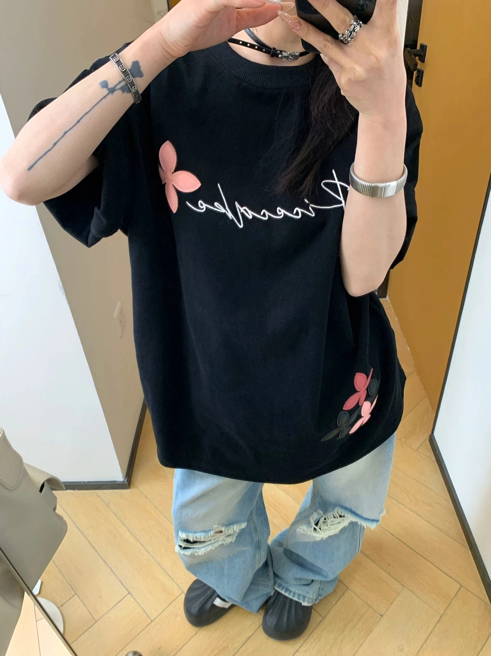 Close-up front view of a black oversized floral graphic T-shirt showing the crew neckline, cursive chest lettering, rolled sleeves, and textured flower details