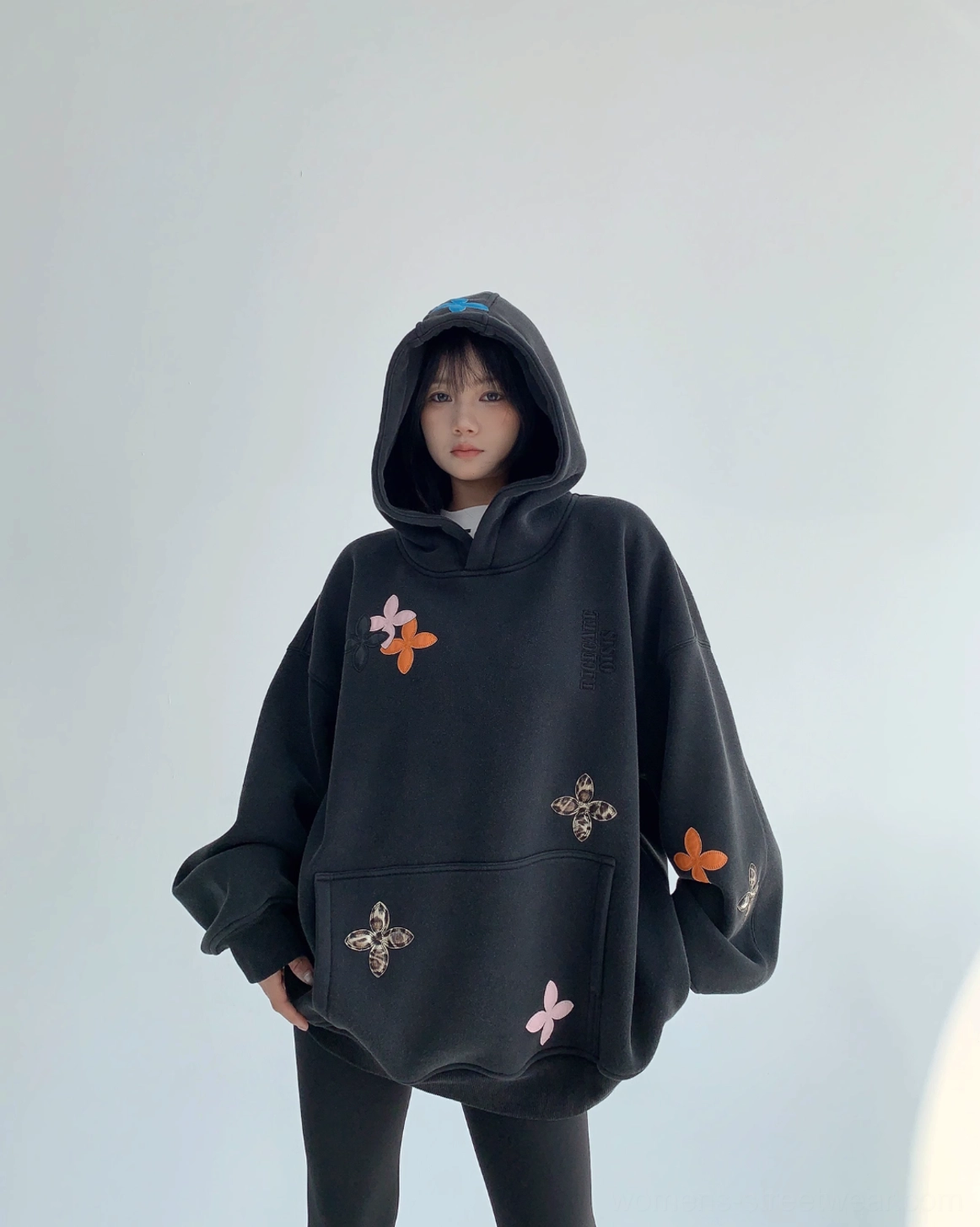 Front three-quarter image of a black oversized floral hoodie with dropped shoulders, colorful flower appliqués, and dramatic loose sleeves