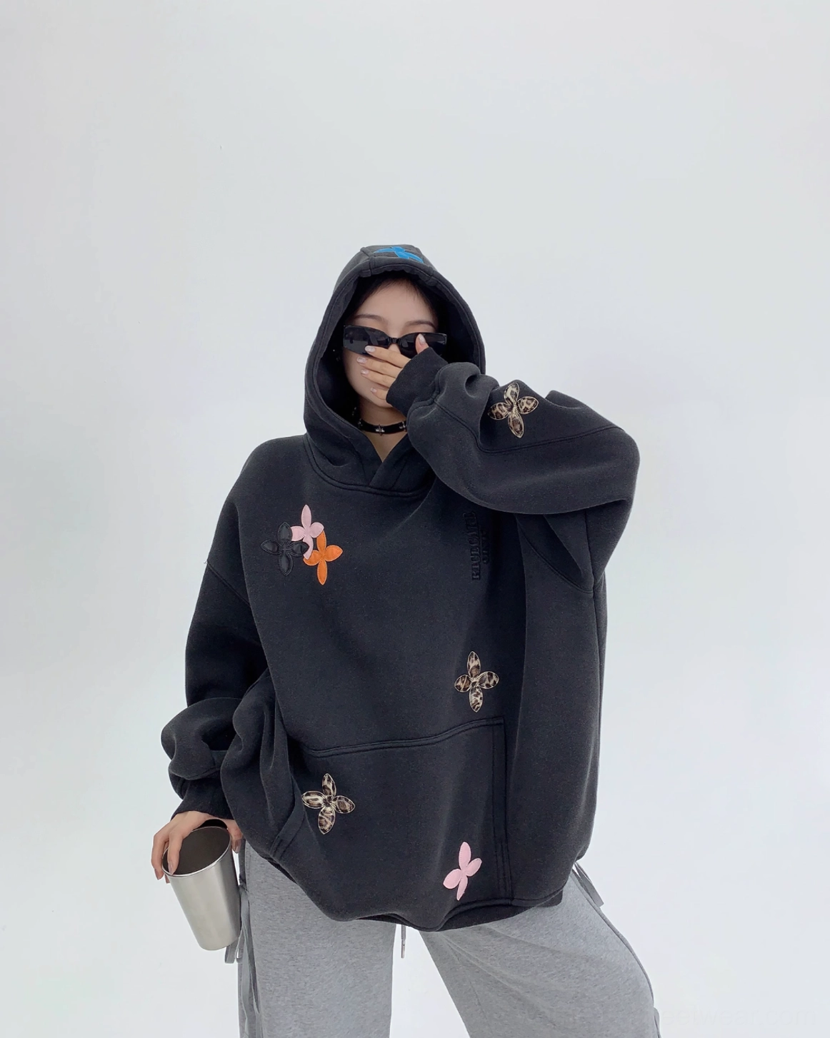 Side three-quarter view of a black oversized graphic hoodie with colorful floral appliqués, long sleeves, large hood, and relaxed Y2K streetwear fit