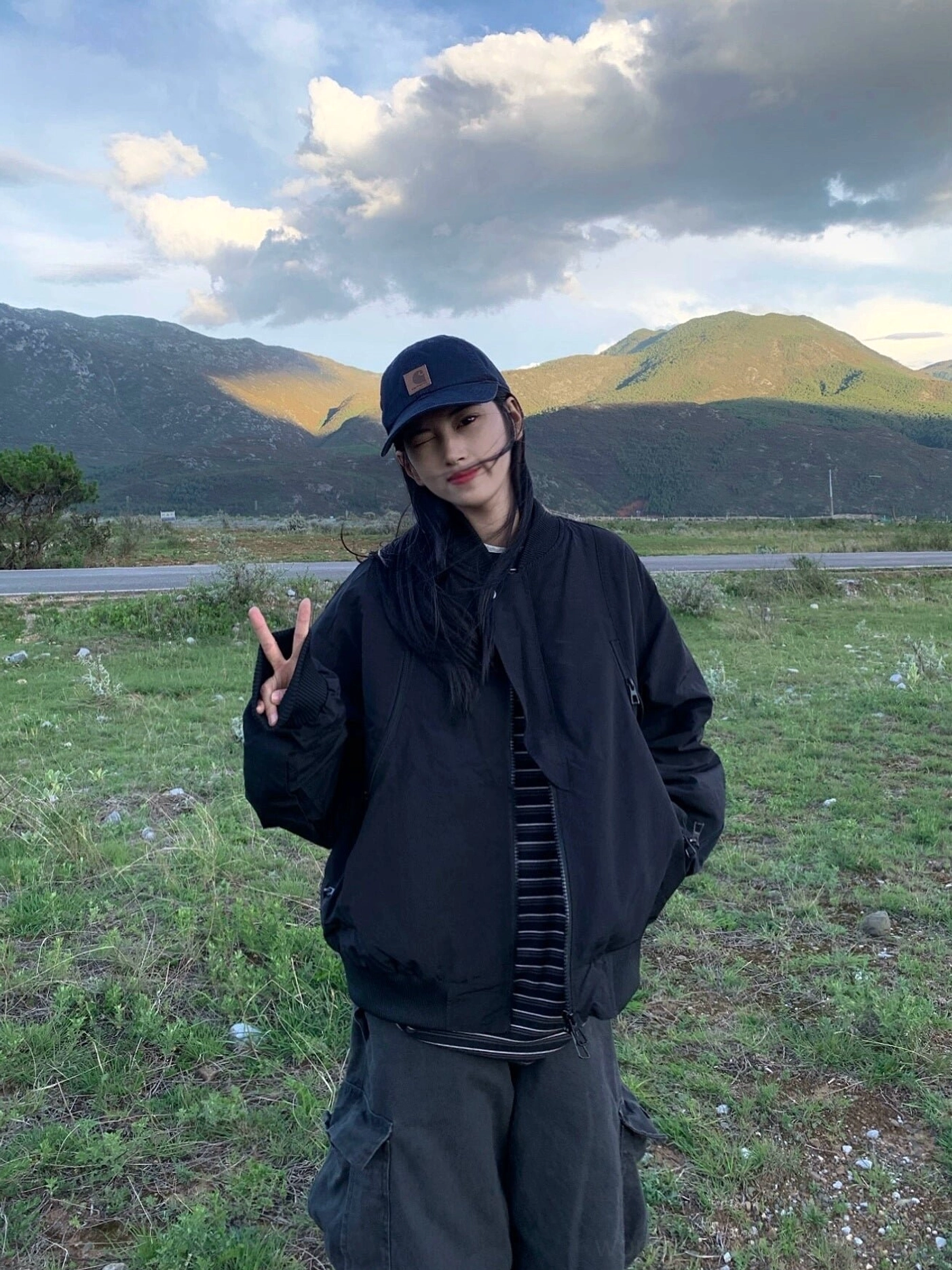Black oversized zip-up flight bomber jacket styled with a striped shirt, cargo pants, and baseball cap on a mountain roadside
