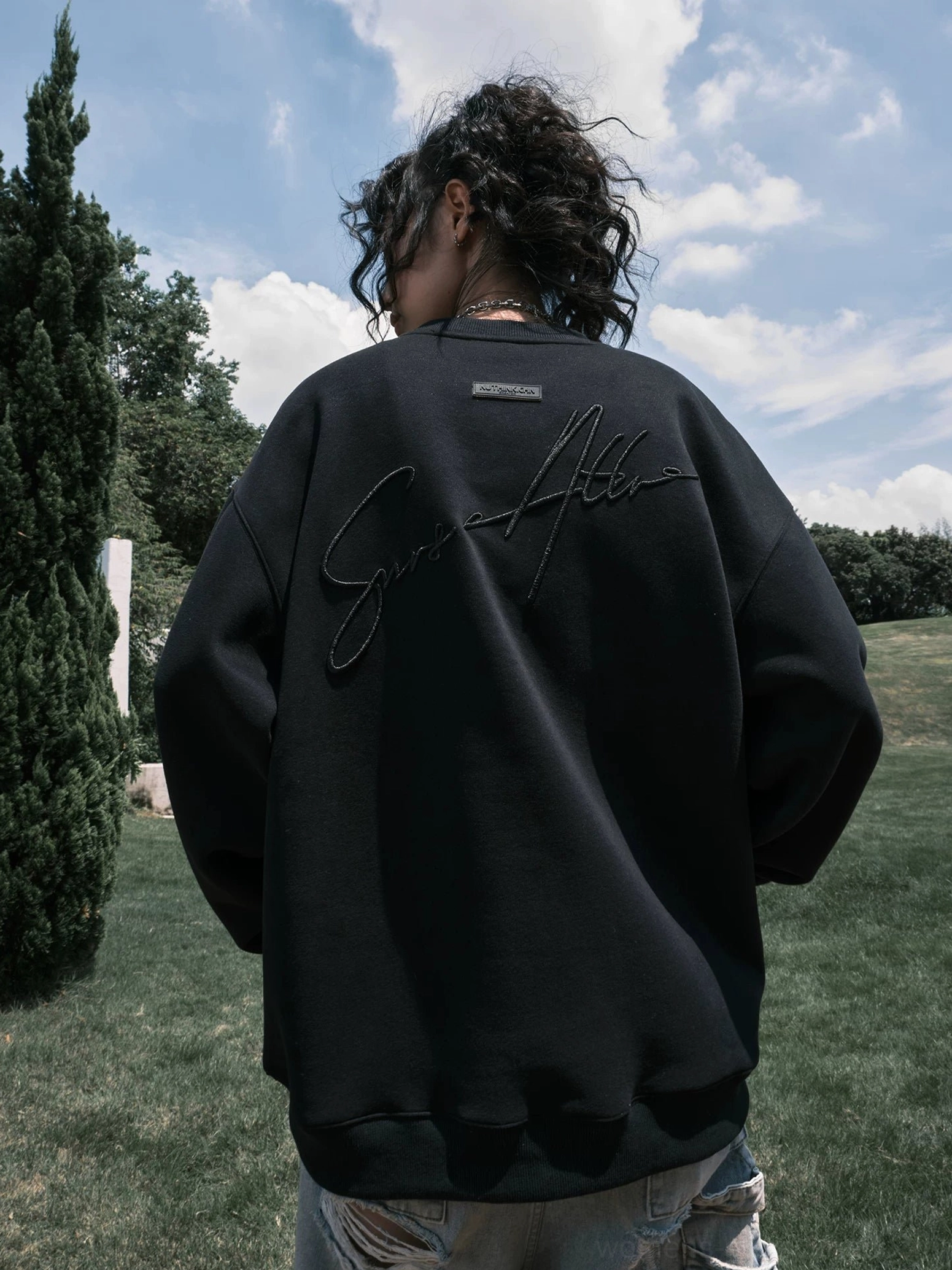 Rear view of a female model in a black oversized streetwear crewneck sweatshirt featuring raised cursive back embroidery and a loose baggy fit