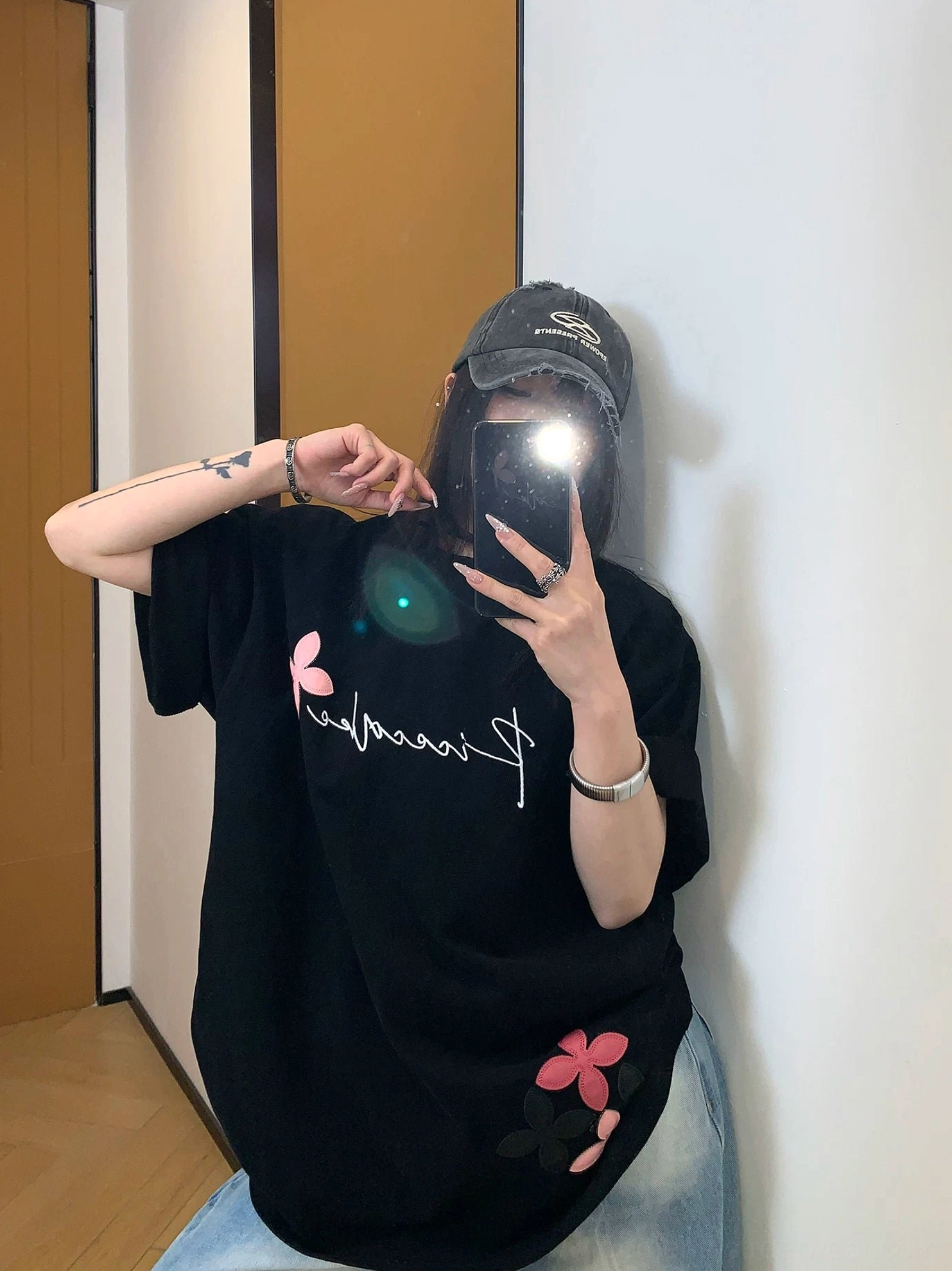 Front mirror selfie showing a black oversized script floral T-shirt with pink flower graphics, rolled short sleeves, and light wash jeans