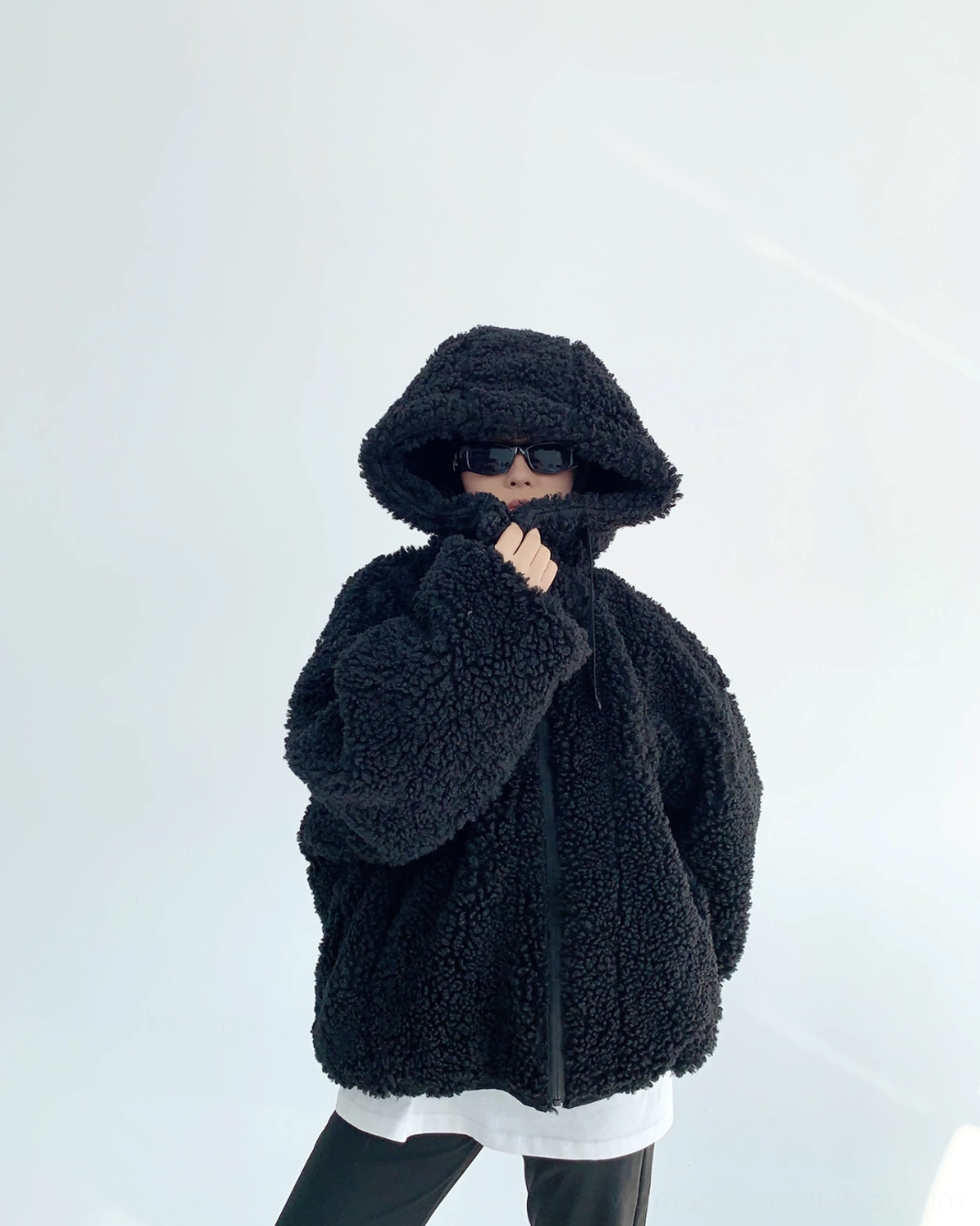 Three-quarter view of woman adjusting the zipper on a black oversized sherpa hoodie with fluffy fleece, hood up, and dropped shoulder sleeves