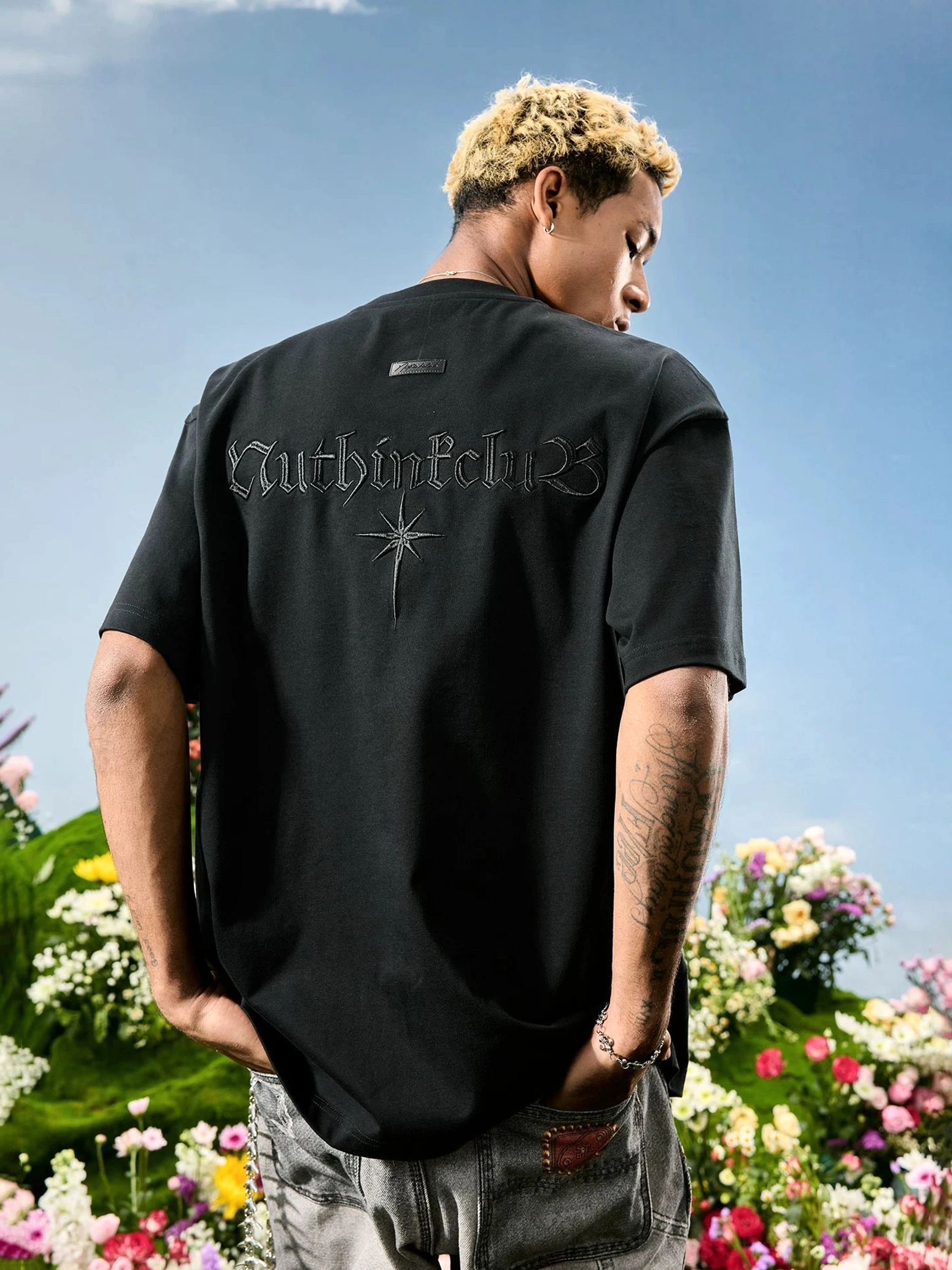 Black oversized unisex streetwear T-shirt on male model, featuring Gothic lettering, starburst back embroidery, and dark monochrome styling