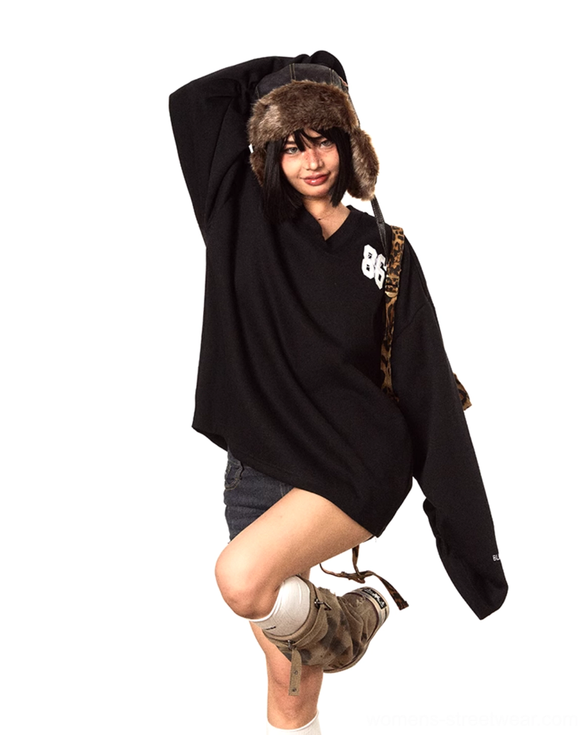 Dynamic three-quarter view of an oversized black V-neck streetwear sweater with embroidered 86 chest detail, loose sleeves, denim shorts, and work boots