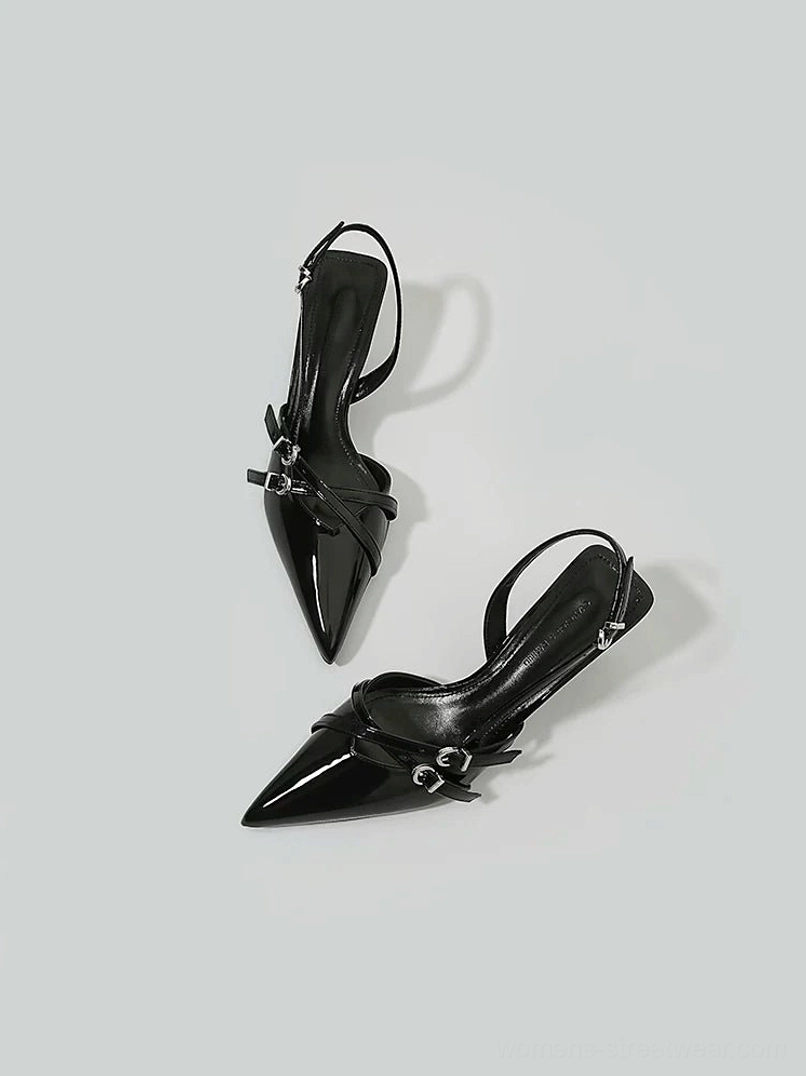 Angled flat lay of black patent slingback heels featuring pointed toe silhouette, crossed straps and metallic buckles
