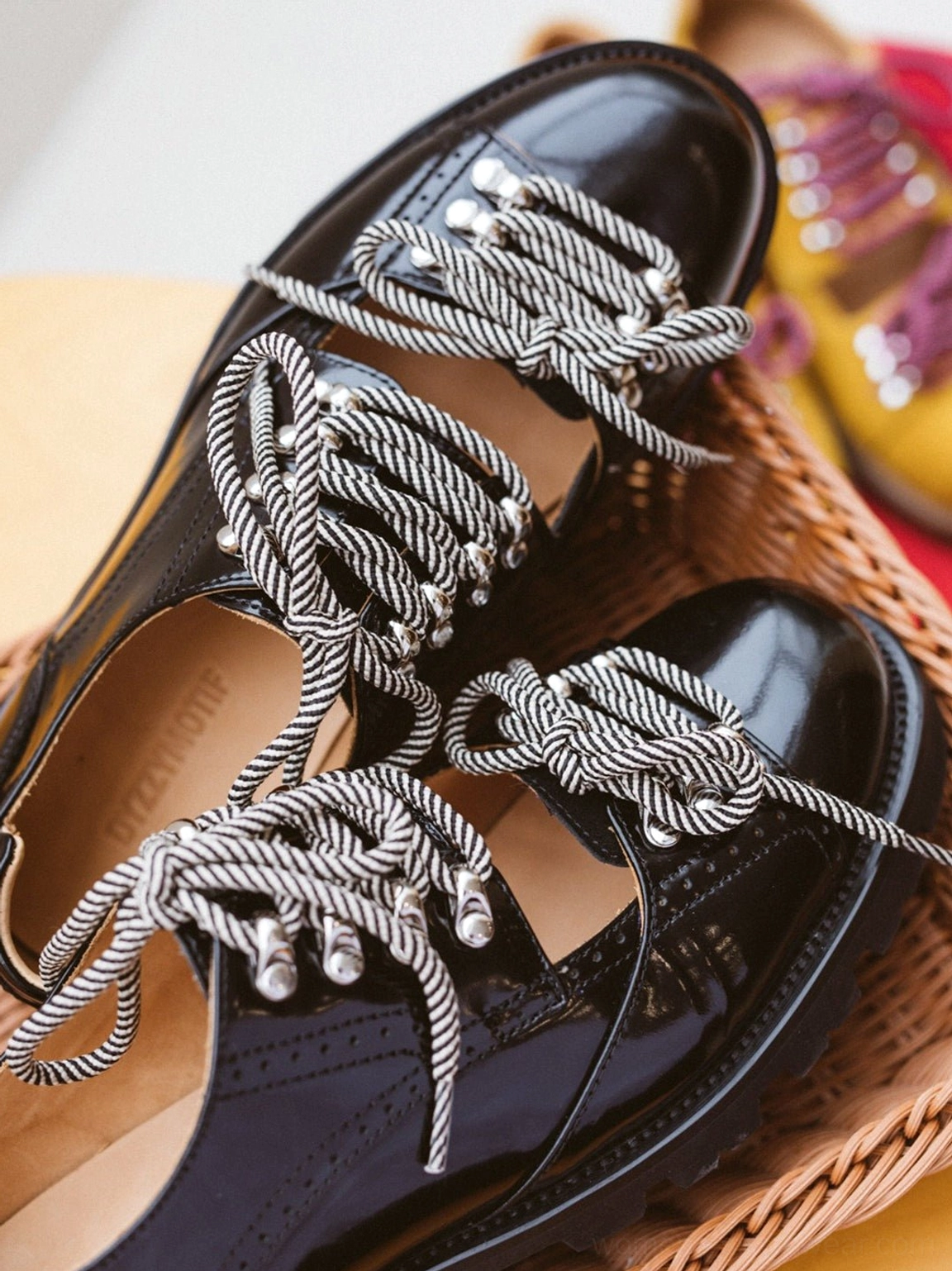 Close-up top-down view of black patent-look cutout workwear derby shoes with gray braided laces, shiny metal eyelets, and mustard yellow pair in the background