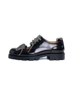 Glossy black patent-look cutout workwear derby shoes with brogue perforations, braided gray laces, metal eyelets, and chunky lug rubber soles on a white background