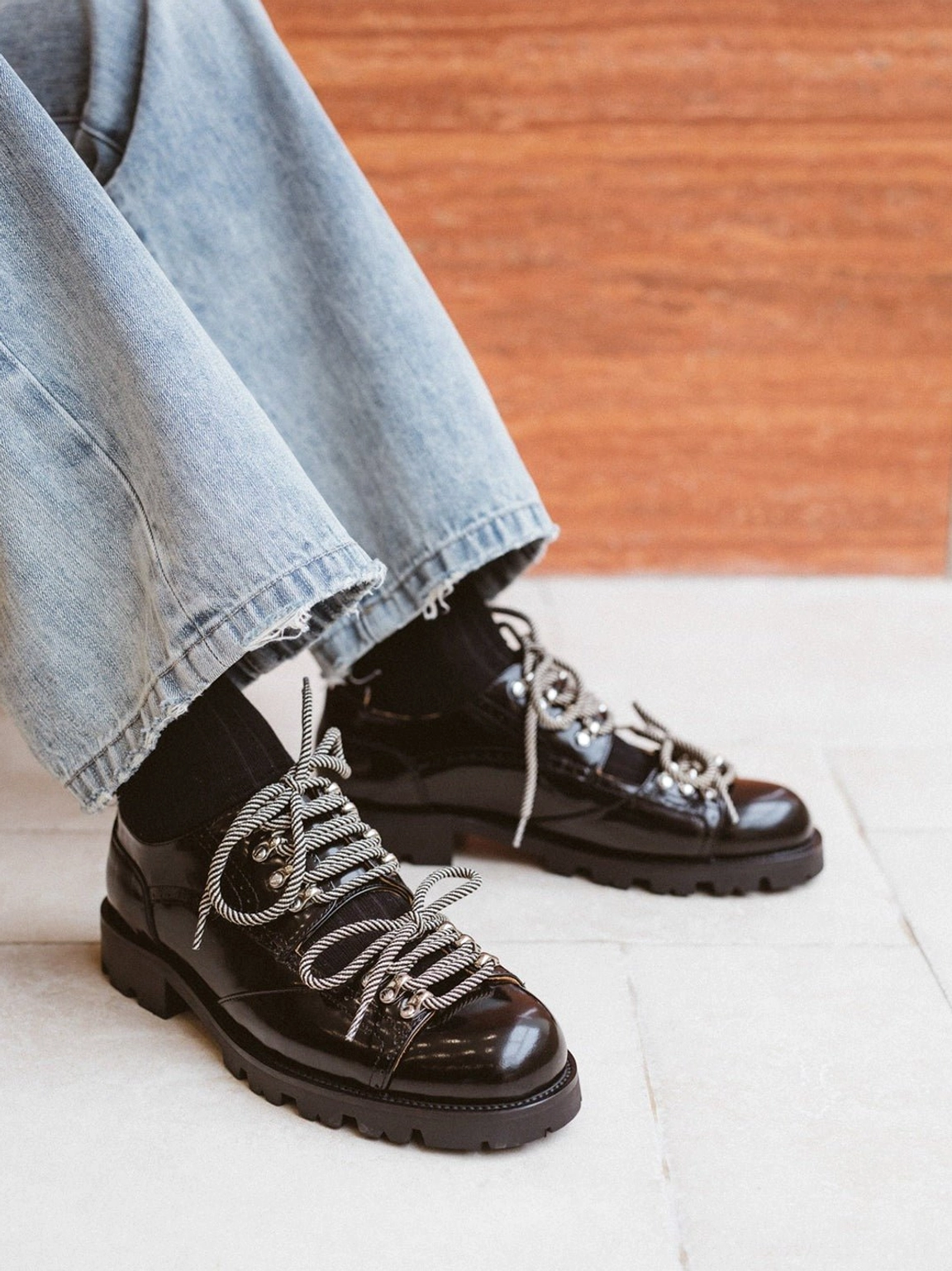 Close-up overhead view of glossy black patent-look derby shoes with silver lace hardware, black socks, light-wash jeans, and thick non-slip lug soles