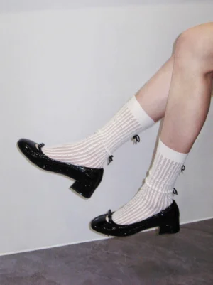 Model wearing black patent leather Mary Jane bow pumps with white ribbed crew socks in a modern indoor setting