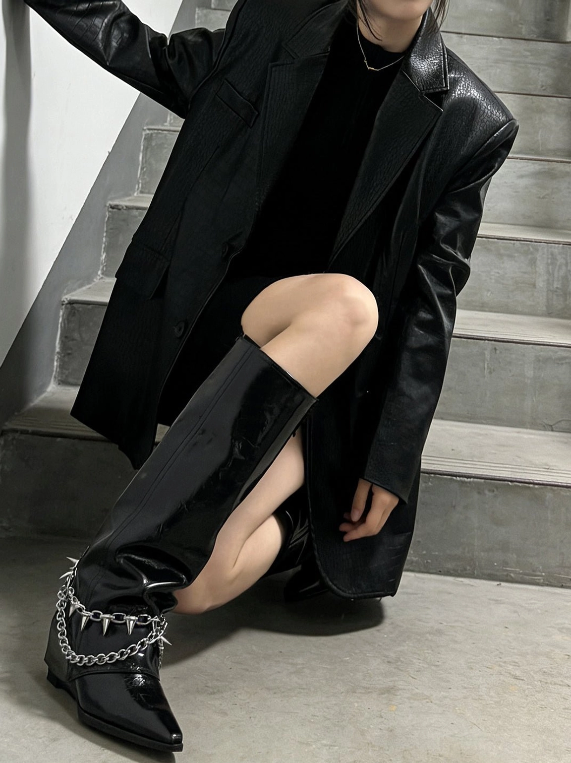 Close low-angle view of black patent spiked chain mid-calf punk wedge boots styled with leather blazer and mini dress