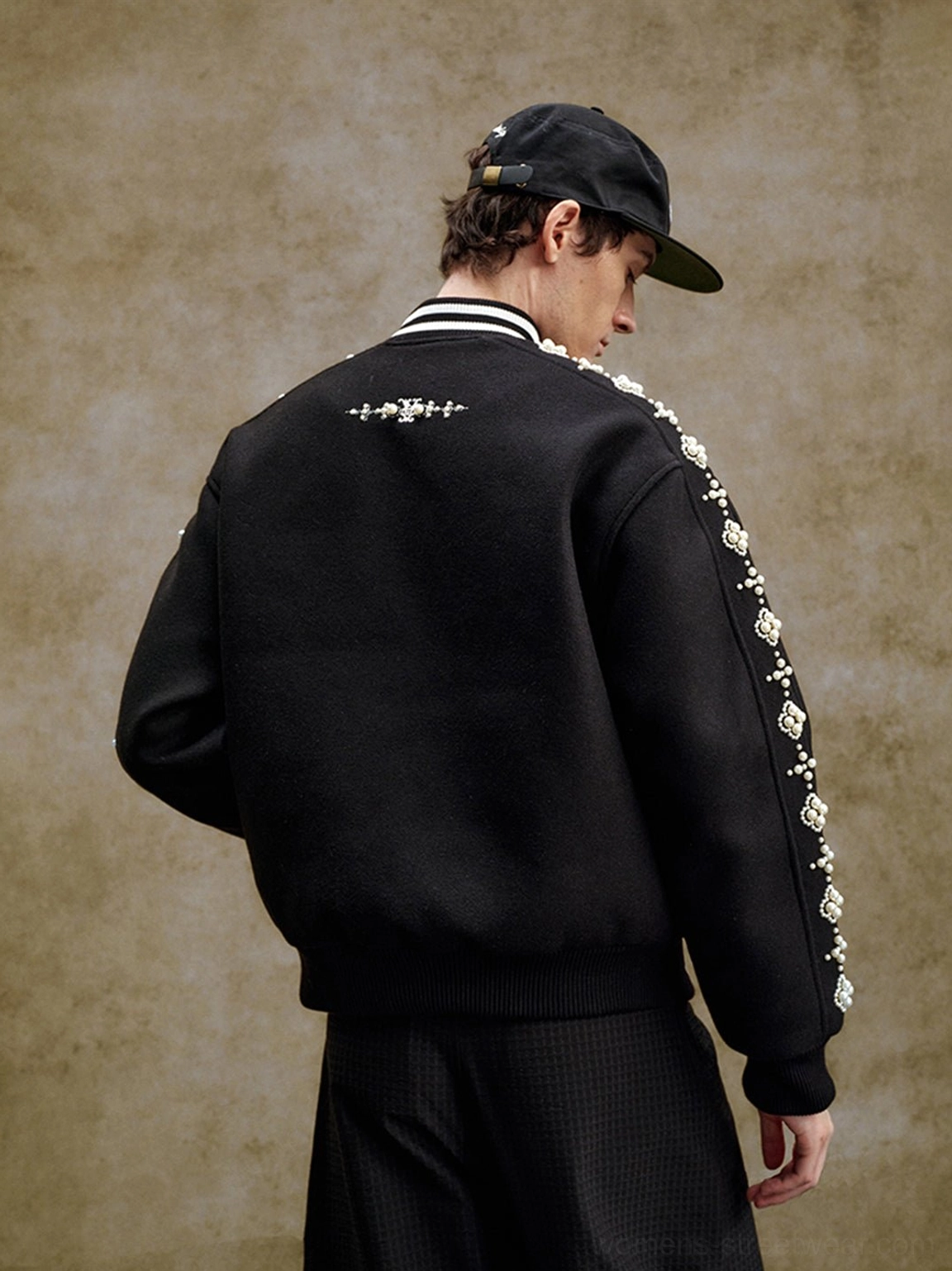 Three-quarter back view of model wearing black pearl beaded varsity jacket with embroidered back motif and ribbed hem