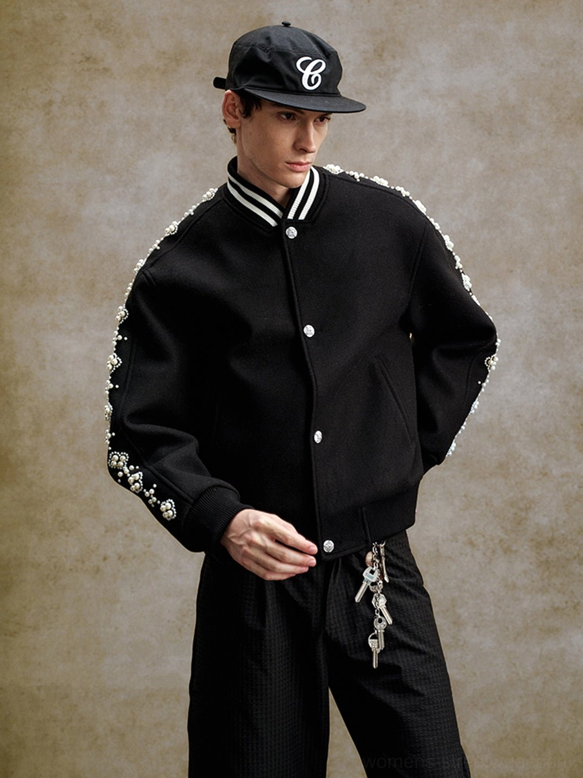 Three-quarter front view of black pearl beaded varsity baseball jacket styled with cap trousers and chain accessory