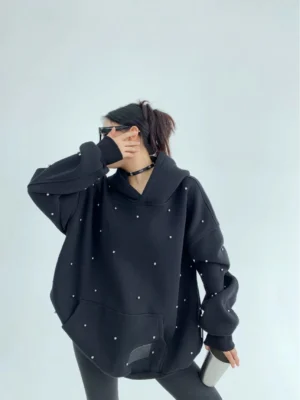Black oversized pearl-studded hoodie shown from a side three-quarter angle with scattered embellishments, oversized sleeves, and hem patch