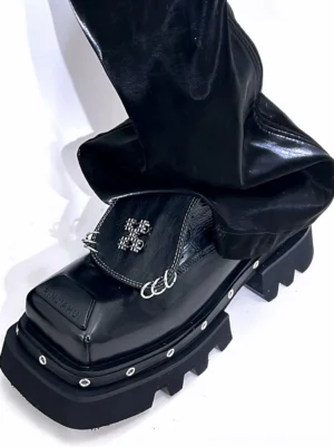 Single black platform Derby shoe worn with shiny black pants, showing silver cross and ring hardware on the decorative flap