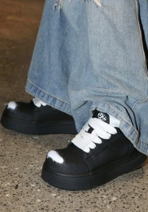 Black chunky platform derby shoes with white spray-painted toe caps styled with loose blue denim jeans in an urban streetwear outfit