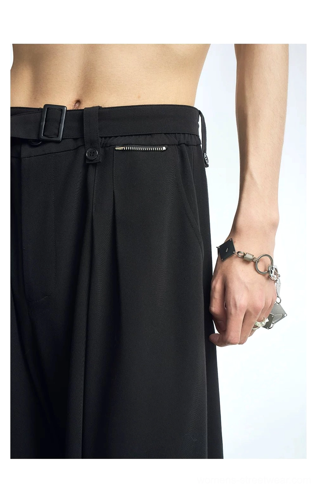 Close-up detail of black pleated streetwear pants waist with adjustable belt loops, zipper pocket, matte suiting fabric, and silver accessory styling