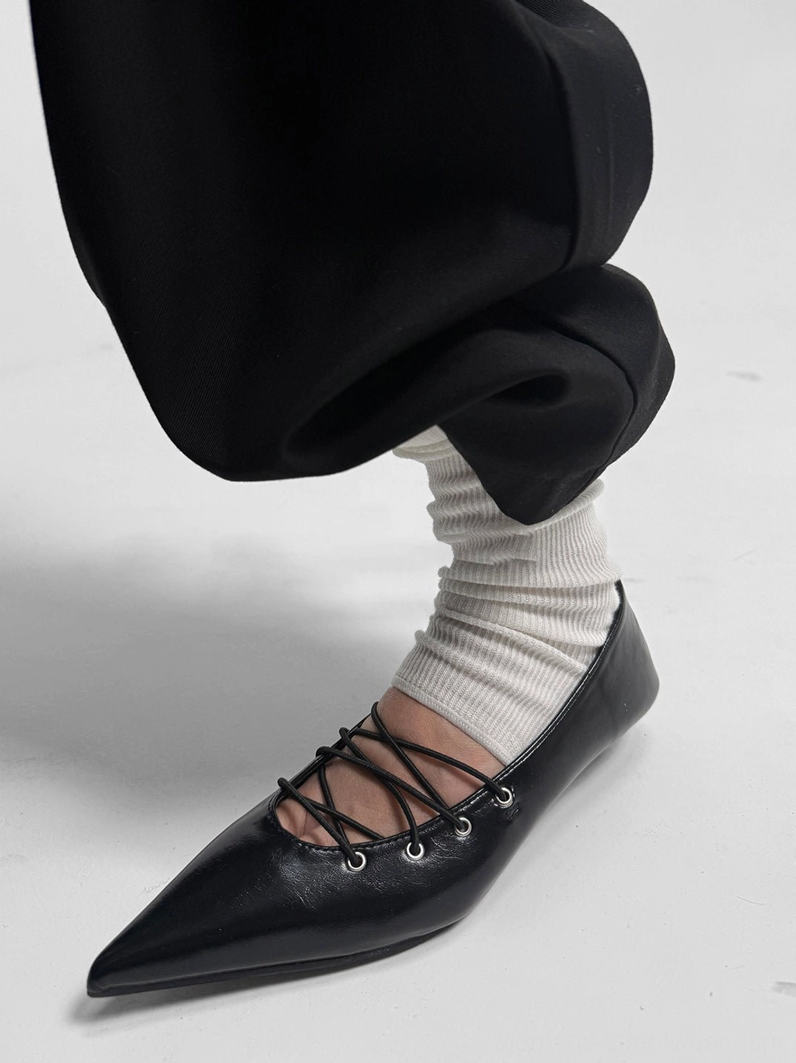 Side view of a black pointed ballet flat featuring crisscross corset lacing, metal eyelets, smooth glossy upper, and low heel