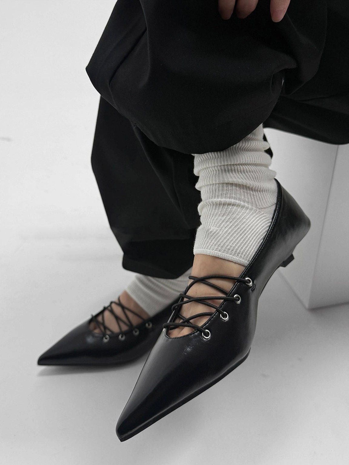 Worn black pointed lace-up ballet flat styled with white slouch socks and loose black trousers in a Korean streetwear outfit