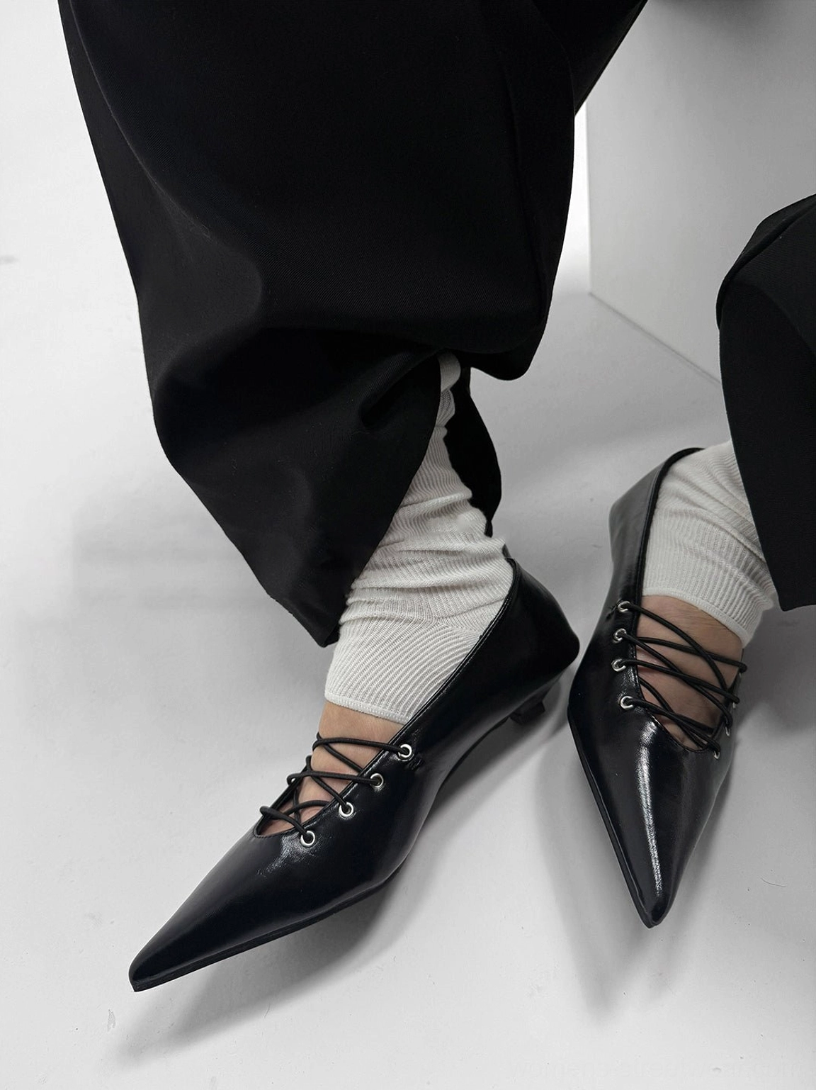 Three-quarter view of black pointed lace-up ballet flats with glossy finish, metal eyelets, and slim kitten heels