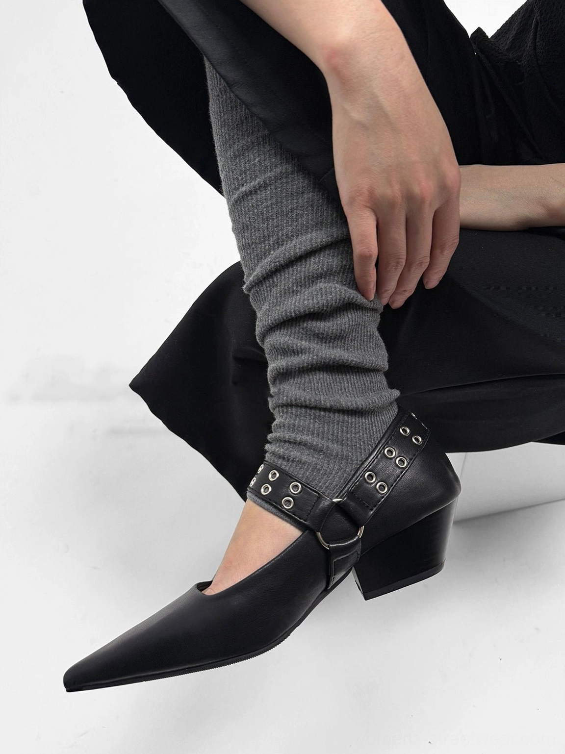 Side view of a seated model wearing black pointed Mary Jane pumps with a sculpted low block heel and hardware strap