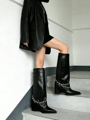 Model wearing black pointed-toe chain riding boots with a knee-high shaft and spike ankle detail styled with a black blazer and mini dress on indoor stairs