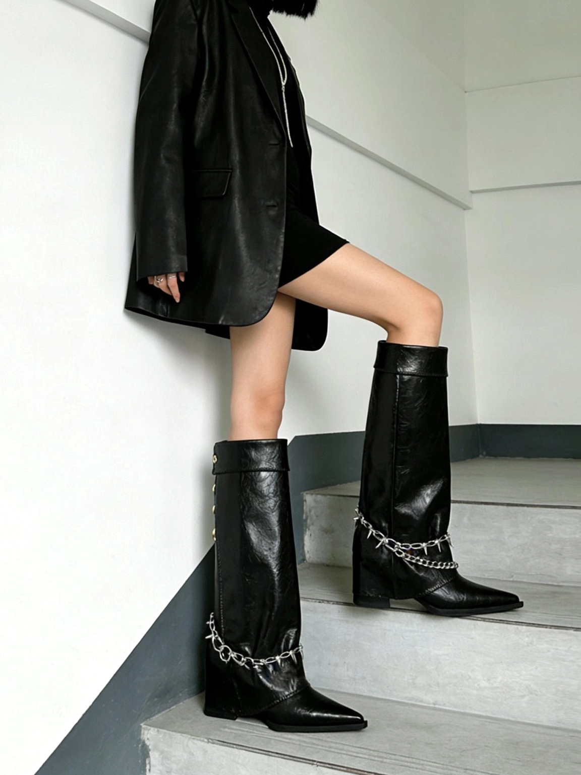 Model wearing black pointed-toe chain riding boots with a knee-high shaft and spike ankle detail styled with a black blazer and mini dress on indoor stairs