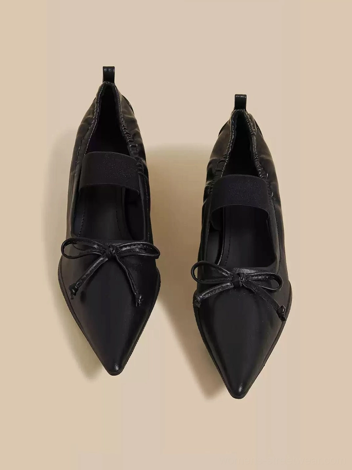 Black pointed-toe Mary Jane mule pair with bow accents and elastic ankle straps on beige background