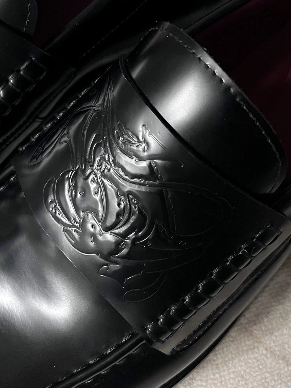 Close-up of a black polished leather loafer with detailed embossed vamp pattern, visible burgundy lambskin lining, and stitched edge trim