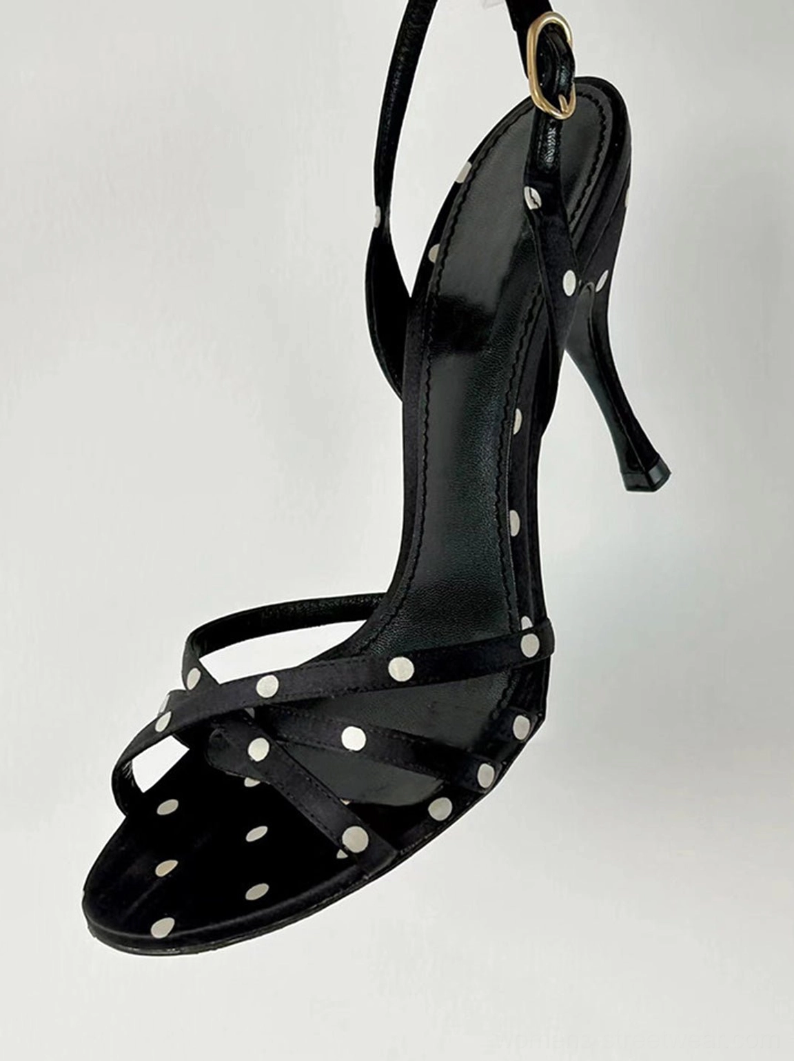 Side profile of a black polka dot strappy stiletto sandal showing the slim heel gold tone buckle and crisscross straps