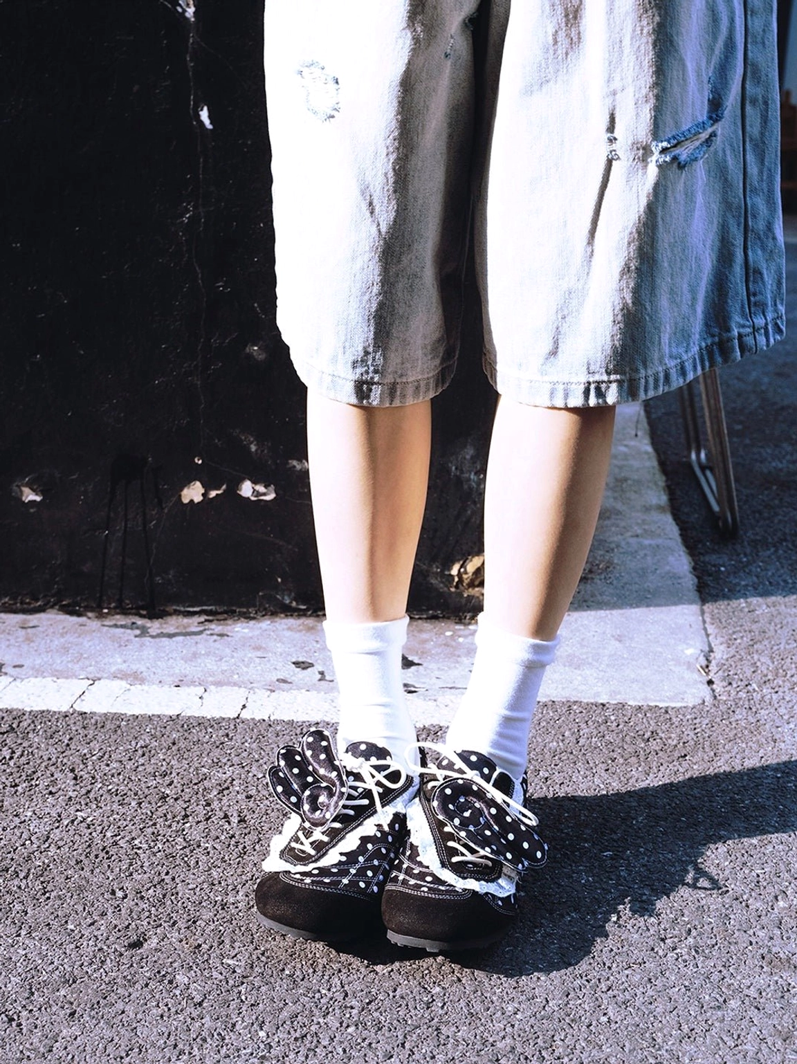 Front low-angle street style view of black polka dot bow ballet flats with white ankle socks and denim shorts on pavement