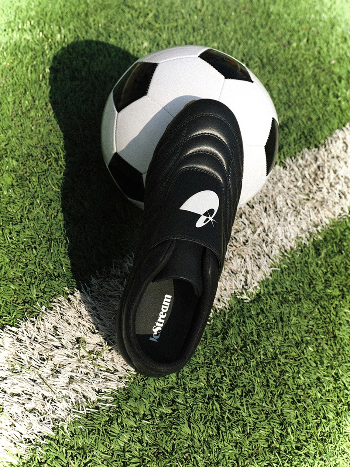 Single black quilted football sneaker beside a soccer ball on artificial turf, showing the strap and gum outsole