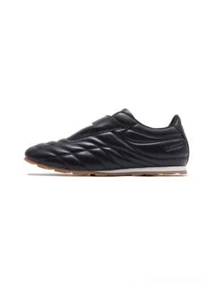 Black quilted leather soccer trainer sneaker with adjustable strap, white tongue patch, and gum rubber outsole on a white studio background