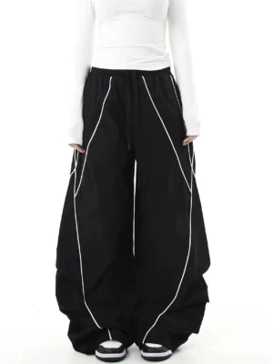 Front full-body view of black reflective curve-leg cargo pants with diagonal piping, oversized wide-leg streetwear silhouette, elastic drawstring waist, and draped lower-leg paneling