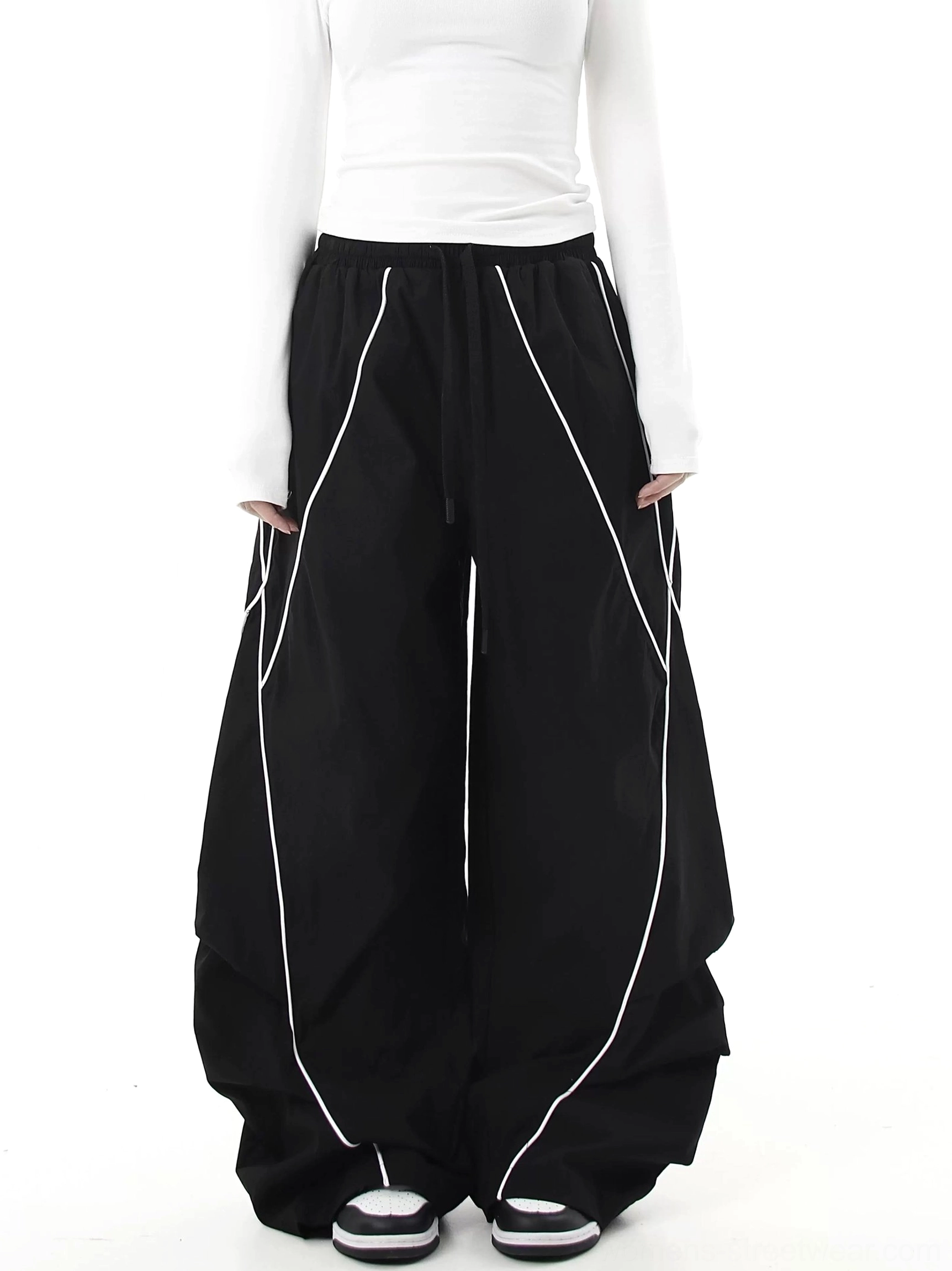 Front full-body view of black reflective curve-leg cargo pants with diagonal piping, oversized wide-leg streetwear silhouette, elastic drawstring waist, and draped lower-leg paneling