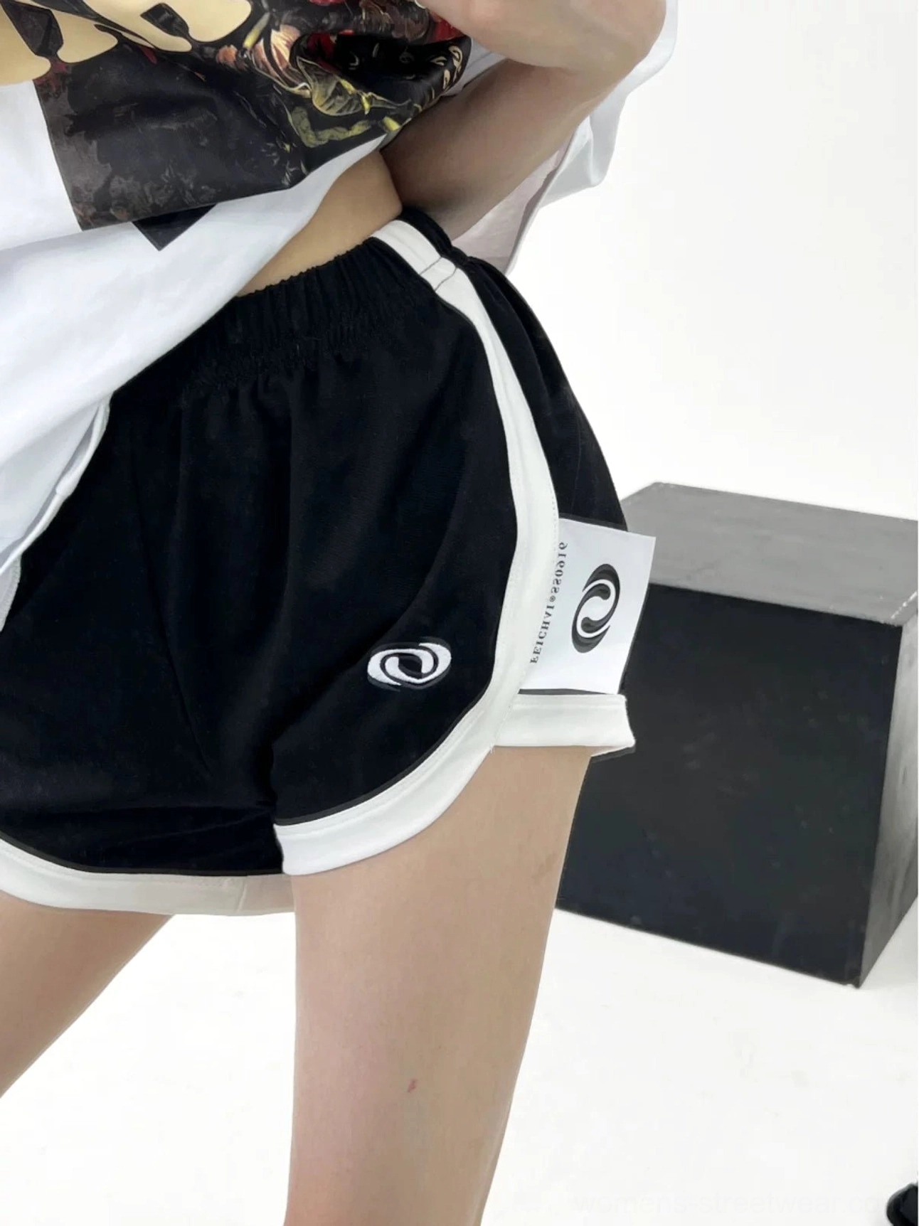 Back close-up of black relaxed fit track shorts showing elastic waistband, white trim, soft gathered shape, and side fabric label