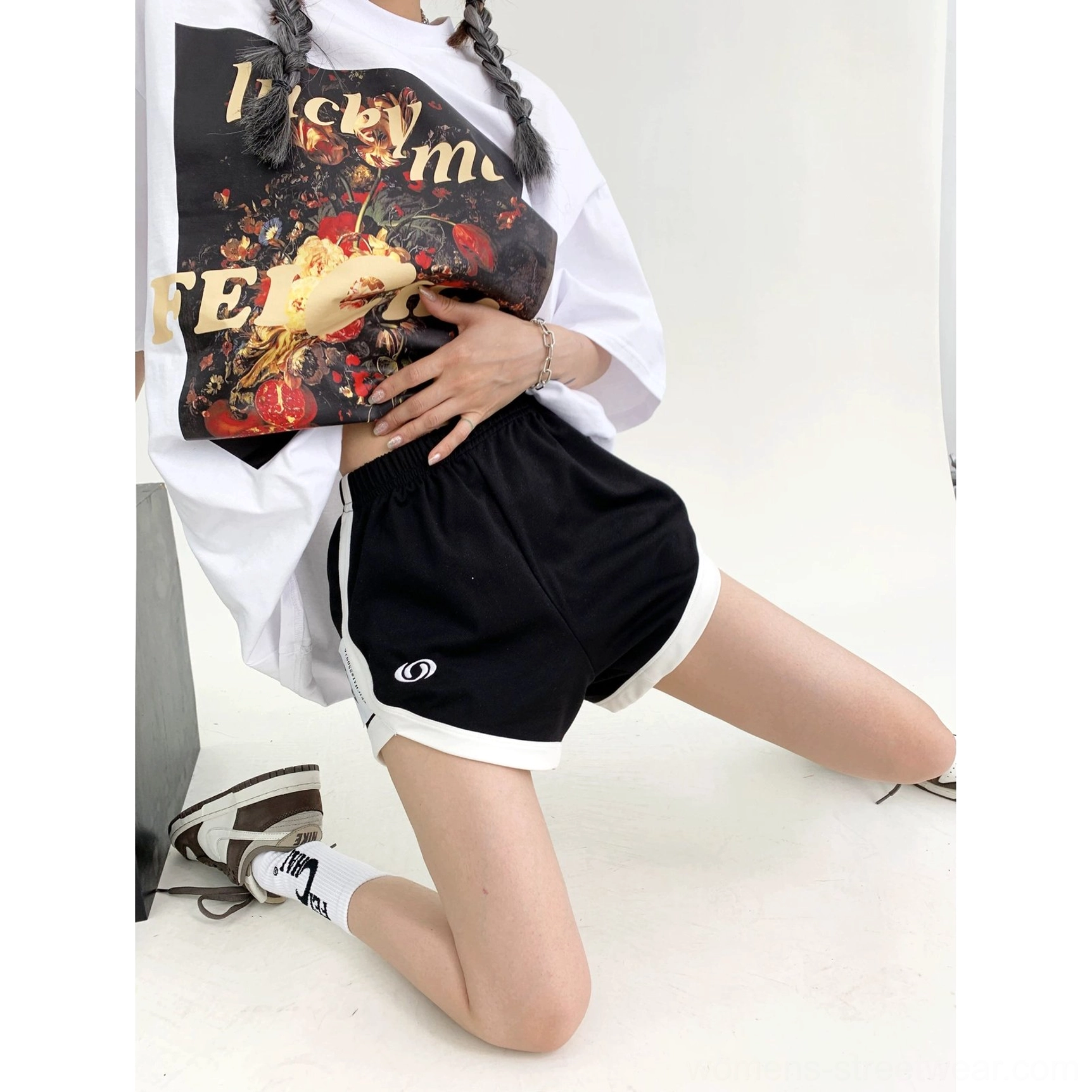 Front kneeling view of relaxed black streetwear shorts with white contrast trim, elastic waist, curved hem, and monochrome styling