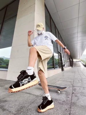 Black retro runner sneakers worn by a male skater in casual streetwear beside a skateboard in an urban corridor