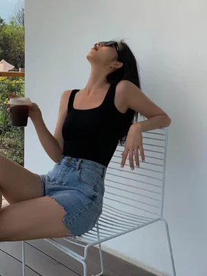 Black square neck ribbed camisole top worn with denim shorts and white metal chair backdrop, showing fitted sleeveless silhouette and wide neckline detail from three-quarter angle