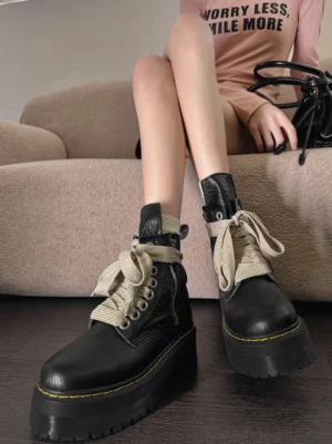 Black chunky platform combat boots with oversized cream ribbon laces, side zippers, padded ankle collars, and yellow contrast sole stitching worn on a model