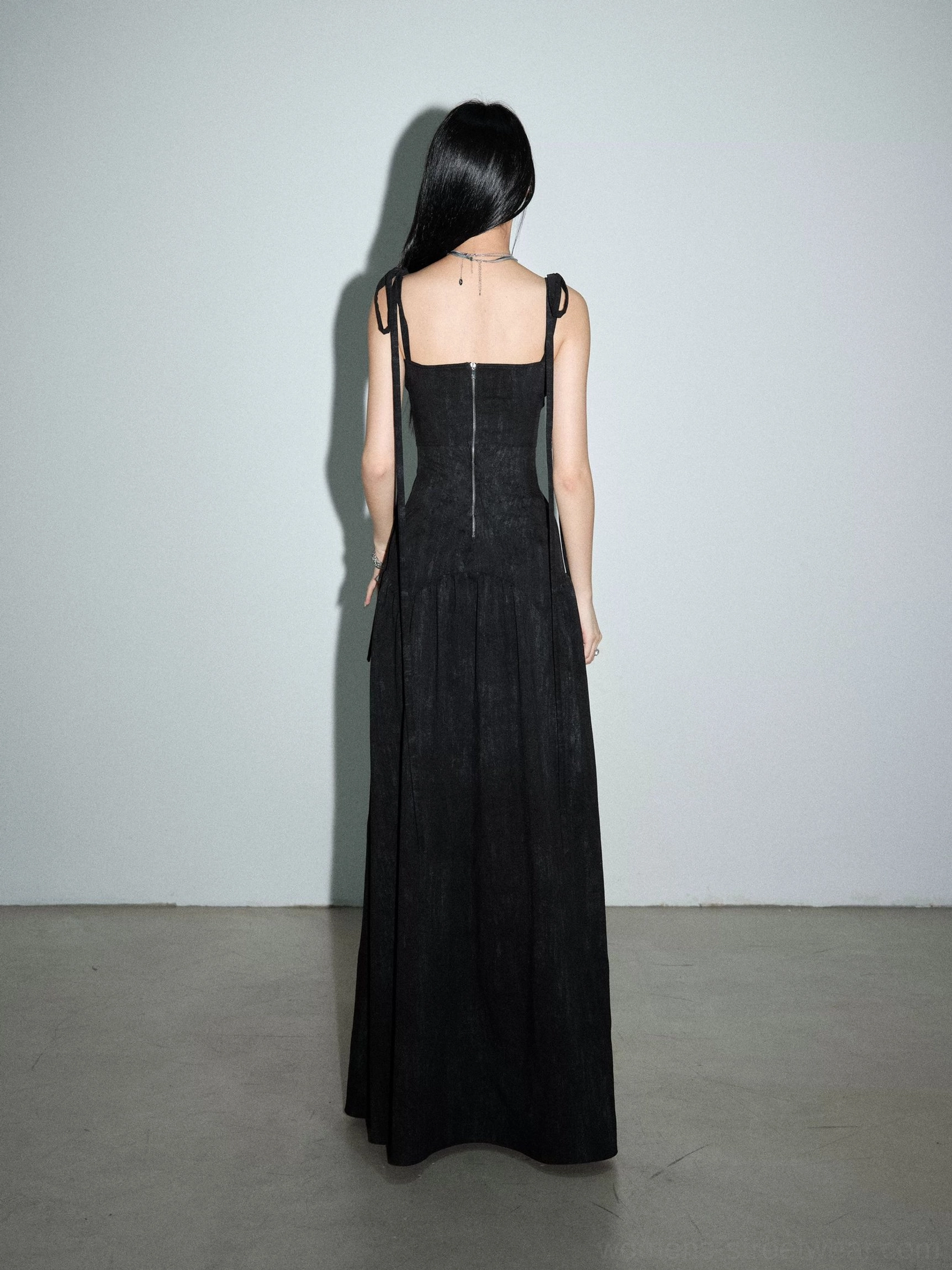 Rear full-body view of a black ribbon-strap maxi dress with exposed back zipper, fitted corset-style back, adjustable ties, and straight floor-length skirt