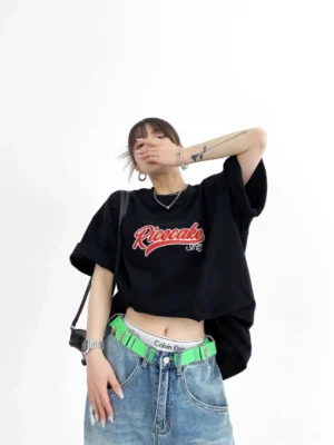 Black Ricecake embroidered oversized varsity streetwear tee front three-quarter view styled with low-rise denim and neon green belt