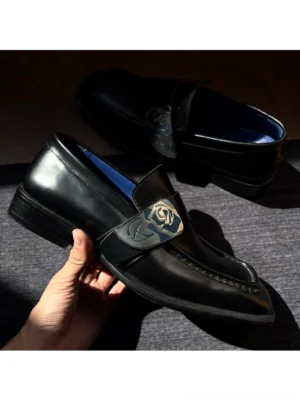 Close-up of black leather square toe loafer with rose shaped metal buckle, blue insole, and polished stitched upper