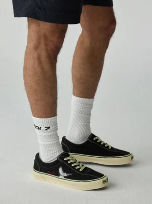 Worn black embroidered low-top skate shoes shown from the rear on a streetwear model wearing casual shorts and crew socks