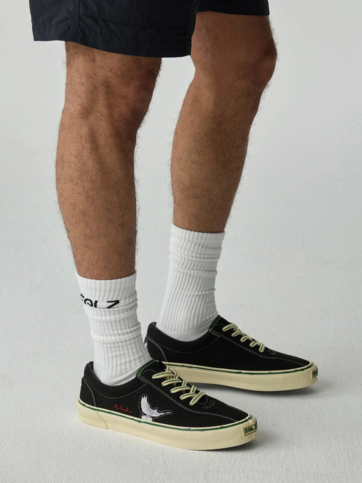 Worn black embroidered low-top skate shoes shown from the rear on a streetwear model wearing casual shorts and crew socks