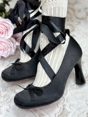Black satin ankle wrap bow pumps styled with white ruffled socks and romantic lace details in a close-up fashion view