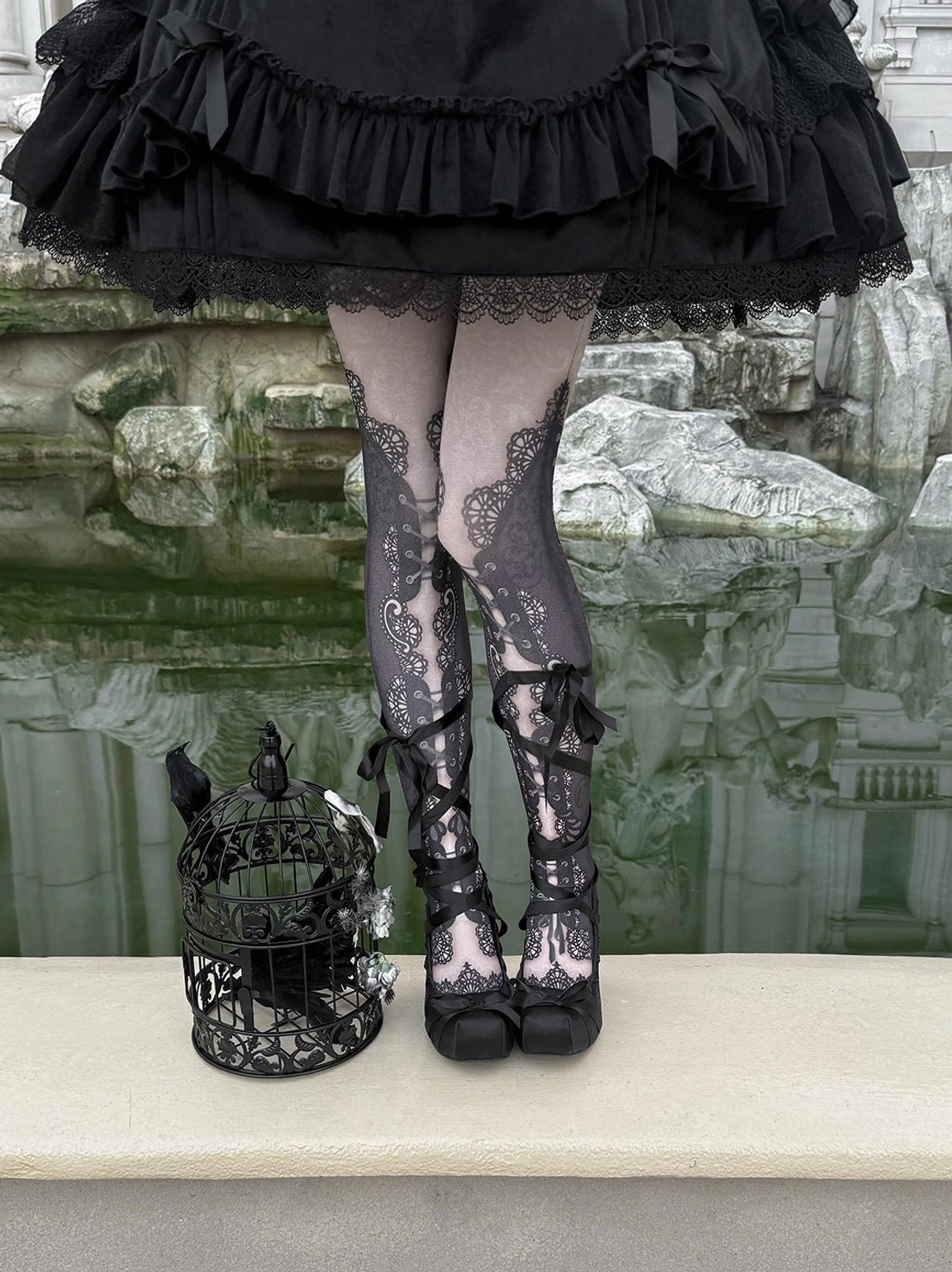 Full lower-body view of black satin lace-up platform pumps with ribbon fasteners, lace tights, and a ruffled gothic Lolita skirt